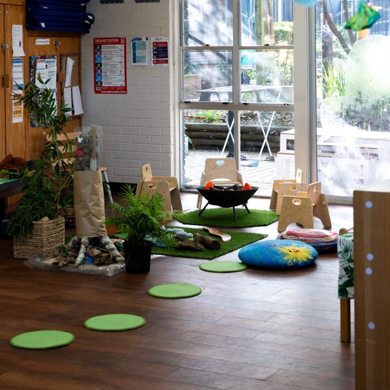 A cozy classroom with wooden chairs around a small table, green circular mats on the floor, and blue and green decorations hanging from the ceiling. Large windows allow natural light. Plants and colorful cushions add a lively touch.